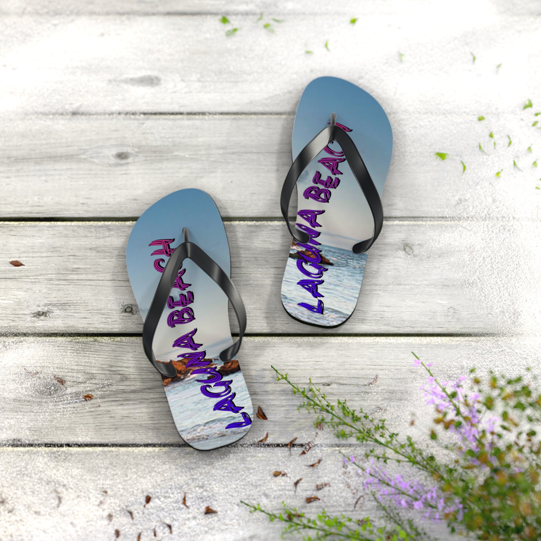 Laguna Beach flip flops on weathered wood with flowers and sand.