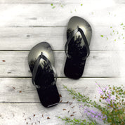 Black flip-flops with a sunset forest scene design on a wooden surface.