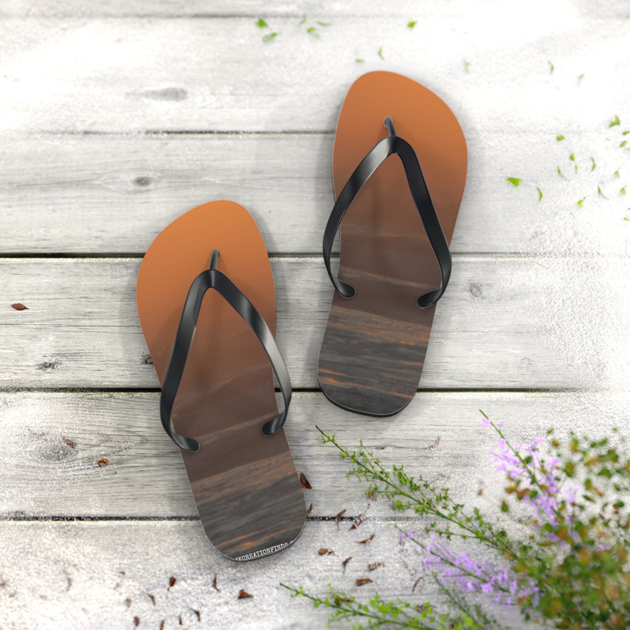Brown flip-flops with sunset design on a wooden surface with scattered petals.