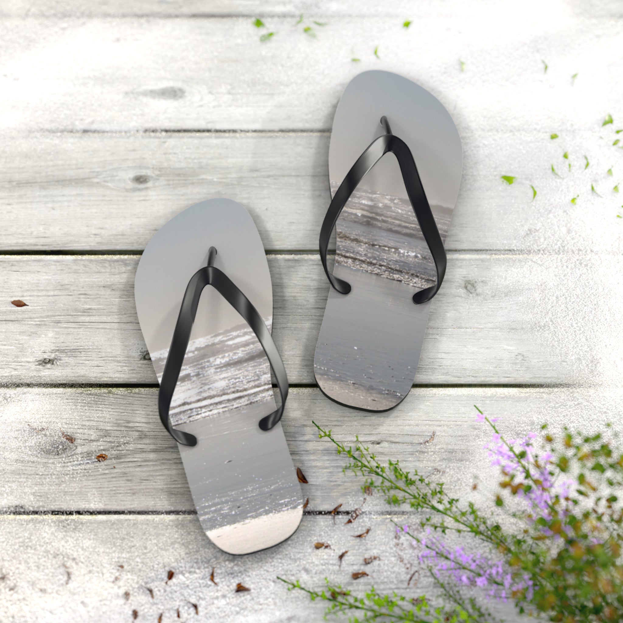 Flip-flops with ocean beach scene on a wooden deck with scattered sand.