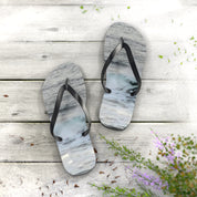 Flip-flops with a wave pattern resting on a wooden surface with sand and flowers.