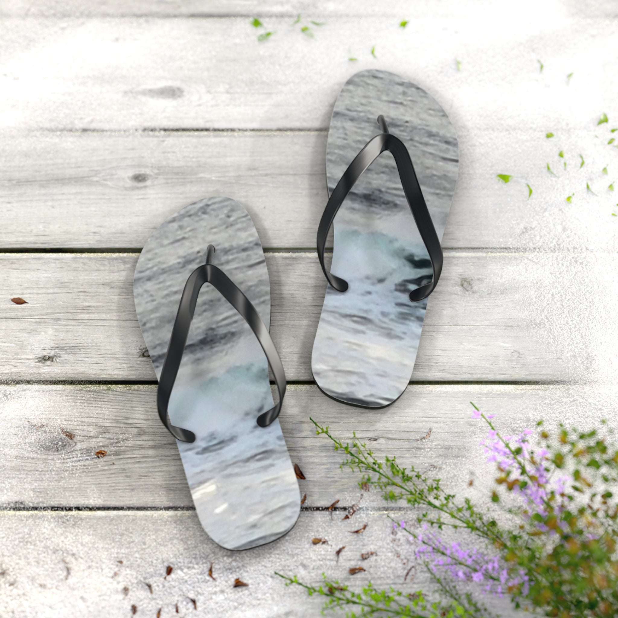 Flip-flops with a wave pattern resting on a wooden surface with sand and flowers.