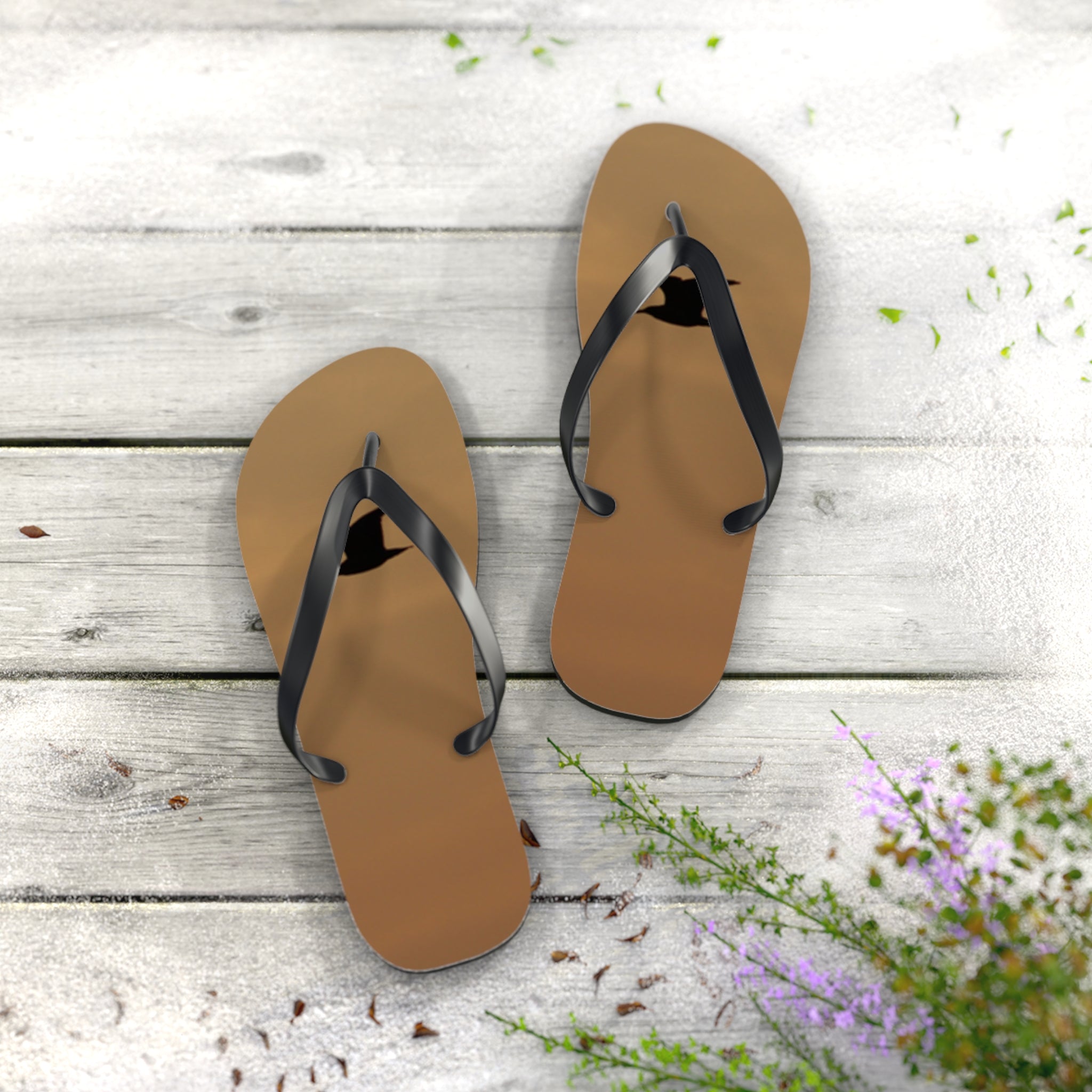 Brown flip-flops with black straps on a weathered wooden surface with small green and purple flowers.