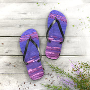 Purple and pink striped flip-flops on a wooden deck with scattered green leaves.